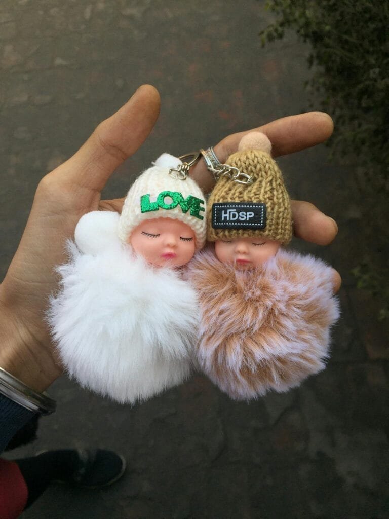 Two fluffy doll keychains held in hand with detailed focus, outdoors in India.
