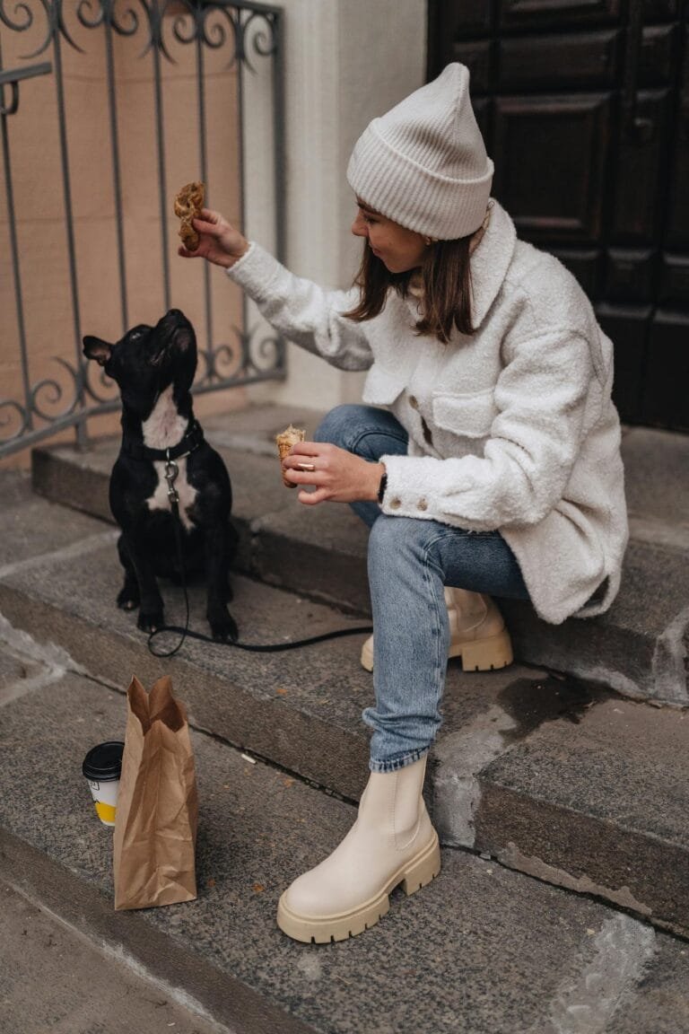 Woman in stylish outfit feeding dog a treat while sitting on concrete steps.